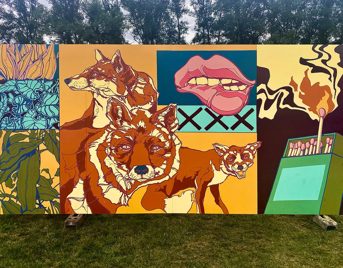 A painted festival hoarding in a field: two foxes, big red lips and a lit match in bold flat colours.
